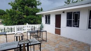 A covered patio area with stone flooring, outdoor furniture, and a white building with windows and a door.