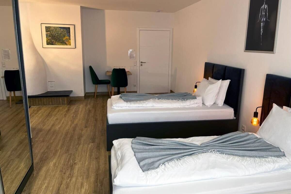 Quarto de hotel com duas camas, piso de madeira, porta, uma cadeira e um quadro na parede.