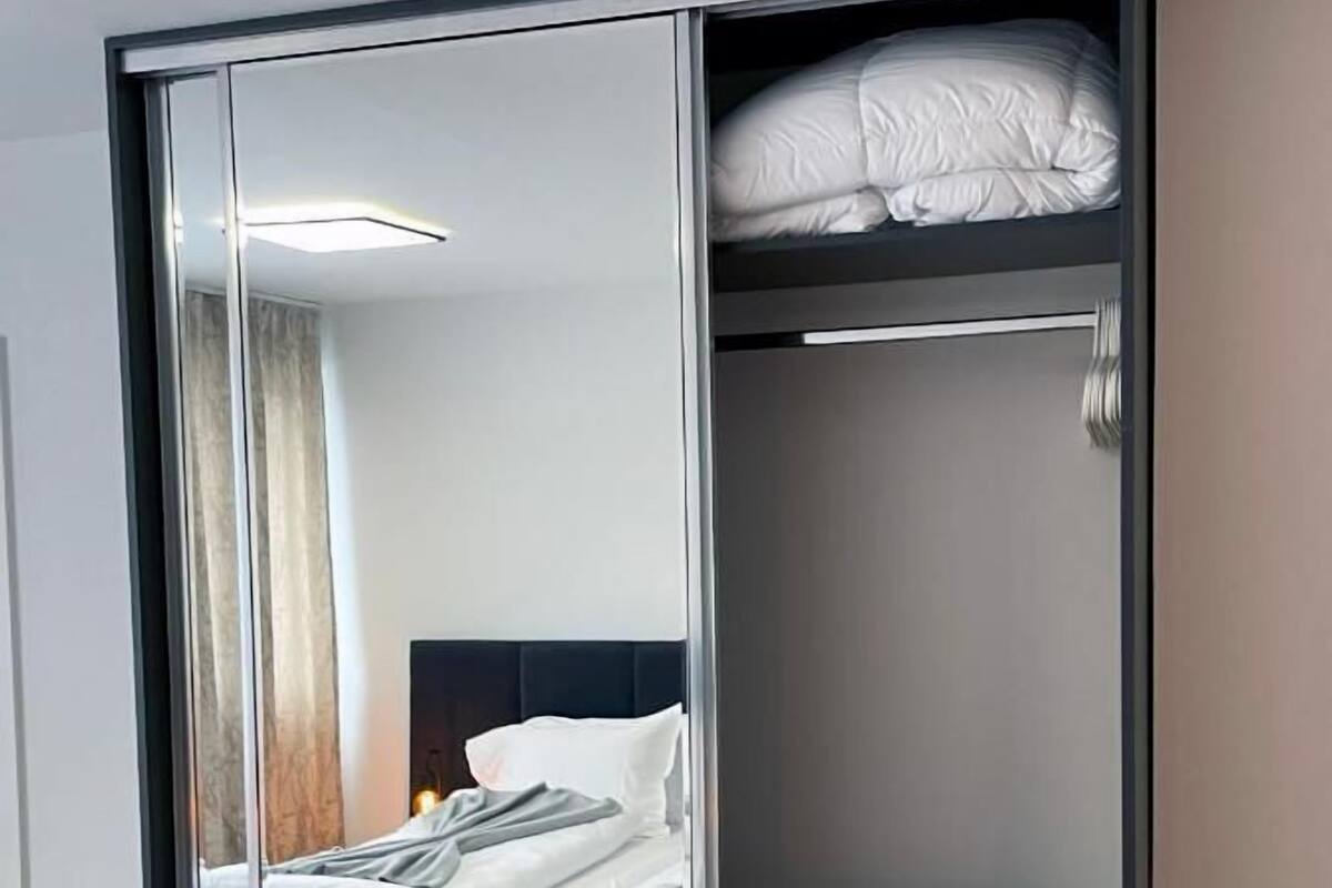 Quarto com guarda-roupa espelhado, cama com roupa de cama branca e chão de madeira.