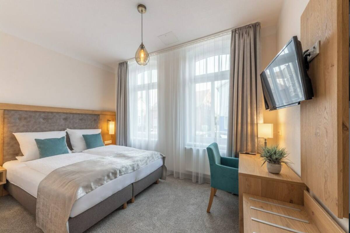 Quarto de hotel com cama, escrivaninha, cadeira, televisão e janela com cortinas.