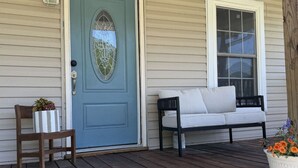 A porch with a blue door, a wooden chair, a white sofa, and a planter with flowers.