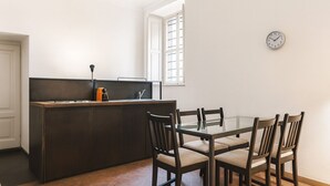 Ett kompakt kök med en mörk träkökö, ett matbord med glasskiva och fyra stolar, en liten orange köksapparat och en väggklocka.