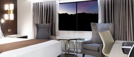 A hotel room with a bed, two armchairs, a small table, and a large window with a view of mountains.