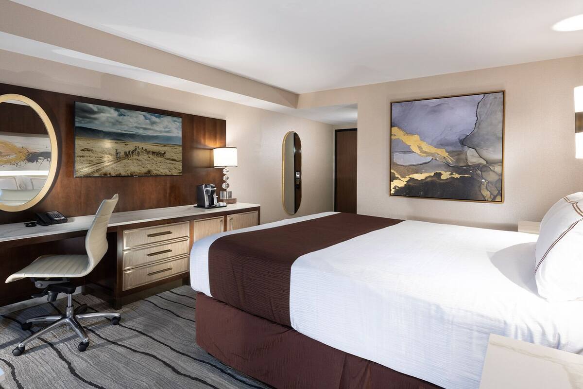 Habitación de hotel con una cama grande, un escritorio con silla, un espejo y dos cuadros en las paredes.