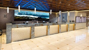 Zona de recepción de un hotel con una gran pantalla digital mostrando un paisaje urbano, un mostrador de mármol y piso de baldosas.