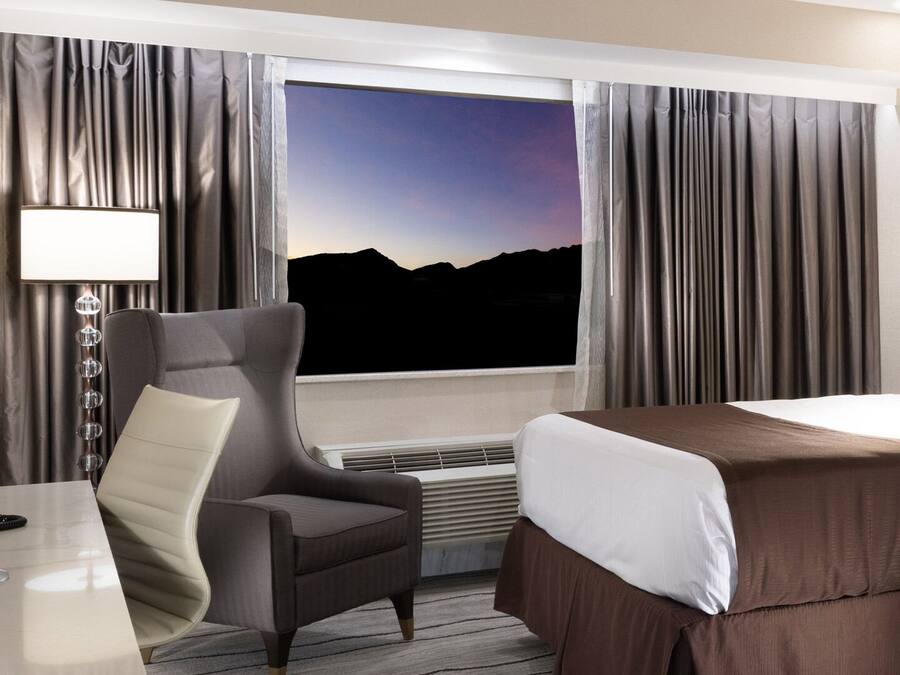 A hotel room with a large bed, a desk with a telephone, a chair, and a window with curtains.