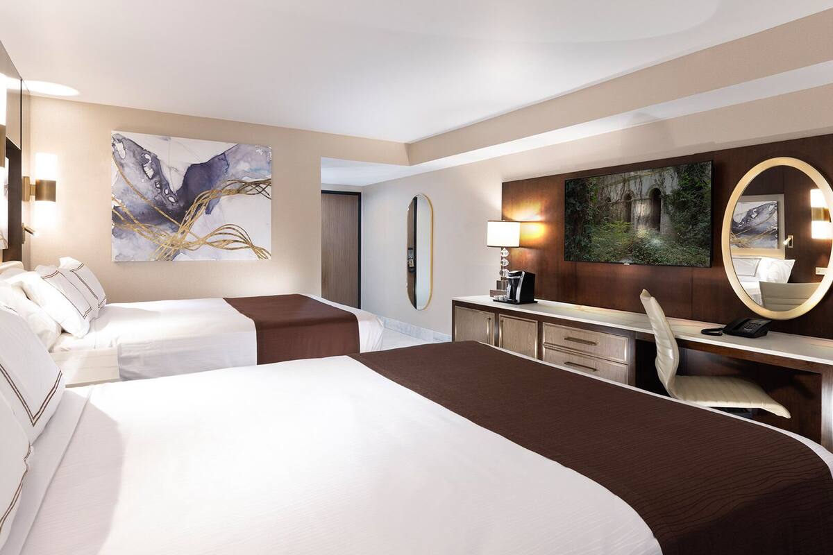 Habitación de hotel con dos camas, un escritorio, una silla, un espejo grande y obras de arte en las paredes.