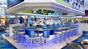 Una zona de bar con varios taburetes y una barra, rodeada de máquinas recreativas y una pantalla grande que muestra deportes.