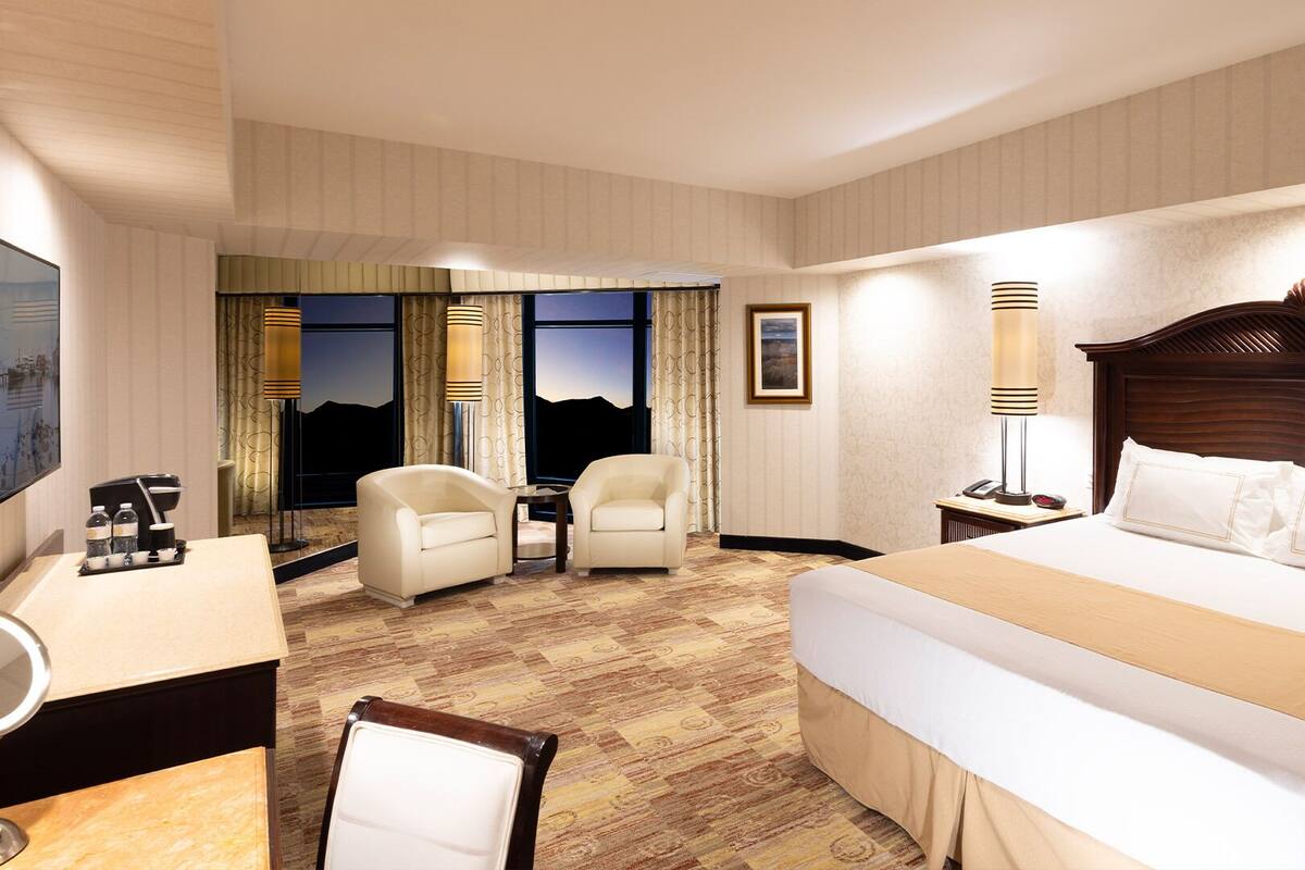 Habitación de hotel con una cama grande, dos sillones, un escritorio con cafetera y vistas a las montañas a través de las ventanas.