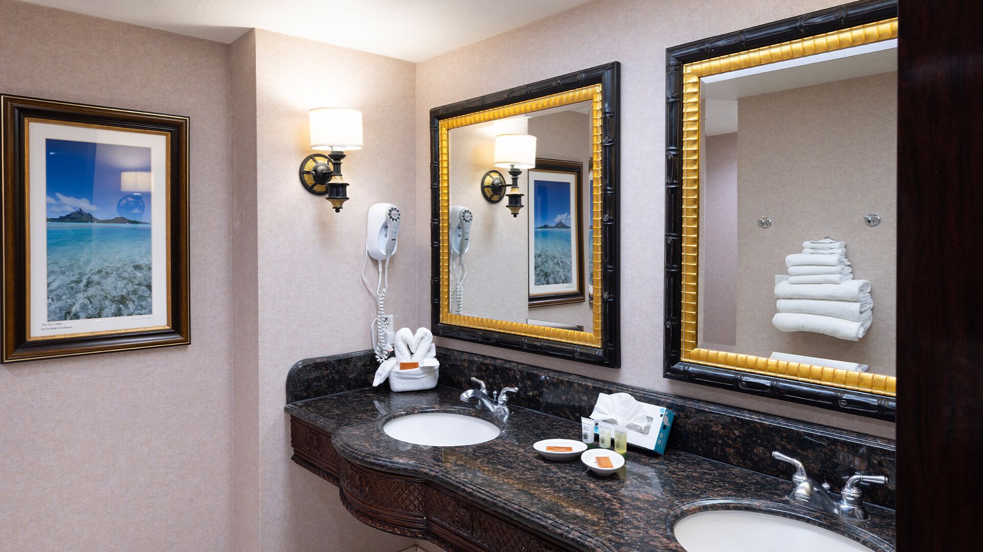 Ein Hotelbad mit zwei Waschbecken, einem großen Spiegel, einem gerahmten Bild an der Wand und einem Handtuchhalter mit Handtüchern.