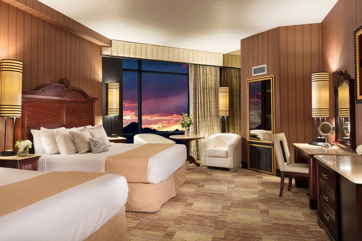 Habitación de hotel con dos camas, un escritorio, una silla y un amplio ventanal con vistas al atardecer.