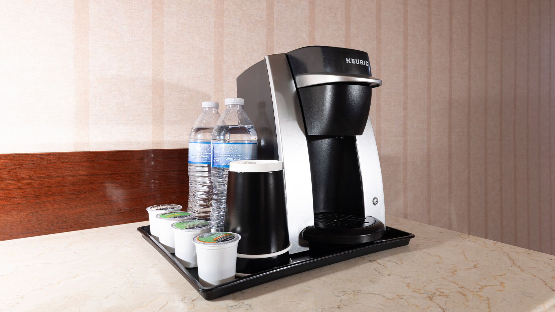 A Keurig coffee maker with a water bottle and two disposable cups on a tray.