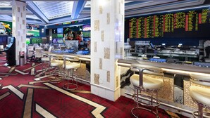 Een drankgelegenheid in een casino met meerdere schermen waarop sportevenementen te zien zijn, barstoelen en een digitaal sportweddenscherm.