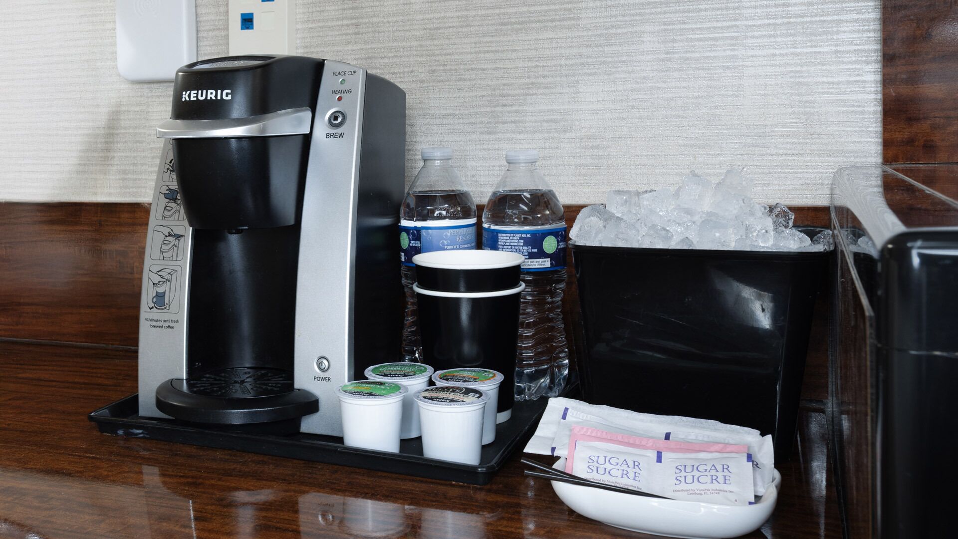 Una cafetera Keurig con un recipiente para goteo, dos botellas de agua, un termo, un tazón de azúcar y un cubo de hielo.