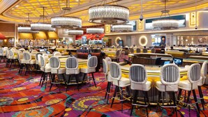 Een ruim casino met meerdere speeltafels en stoelen, een levendig tapijt en weelderige kroonluchters.