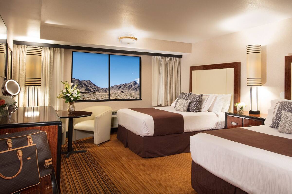 Une chambre d’hôtel avec deux lits, un bureau avec une chaise, une coiffeuse avec un miroir et une grande fenêtre donnant sur la montagne.