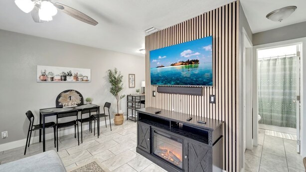 A modern living room with a flat-screen TV mounted on a wooden panel wall, a fireplace, a dining area with a table and chairs, and a ceiling fan.