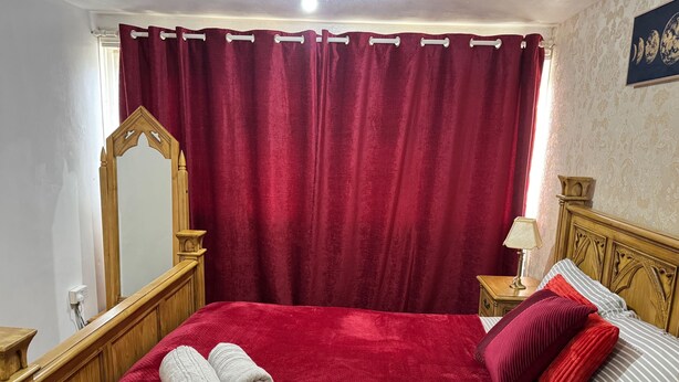 Una cama bien tendida con una manta roja y dos toallas enrolladas, un cabecero de madera, una mesita de noche con una lámpara y un espejo grande en la pared.