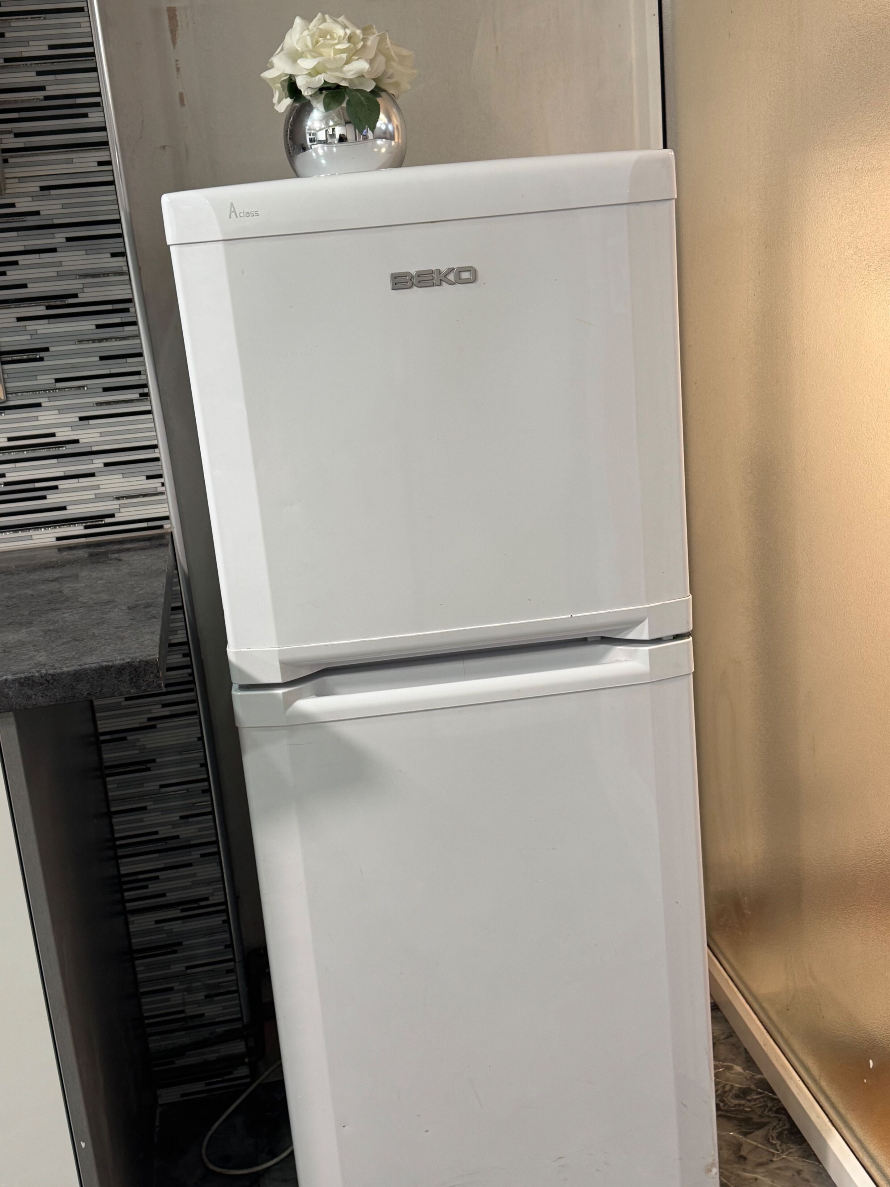 Un réfrigérateur BEKO blanc dans une cuisine avec un mur arrière à motifs et un mur de couleur beige.