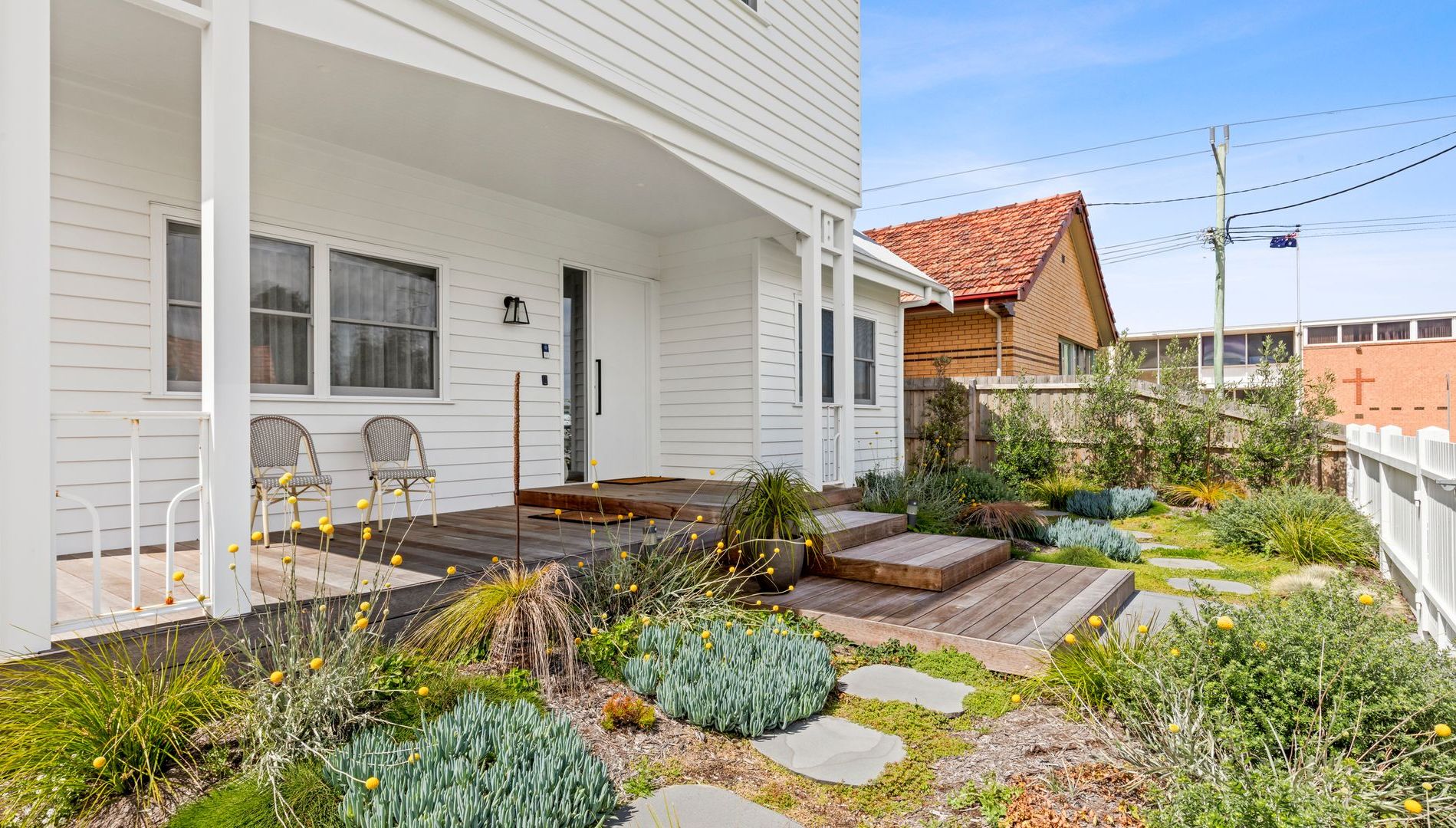 A white house with a covered porch, a brick building in the background, and a landscaped garden with various plants and a wooden deck.