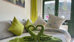 A cozy room with a sofa adorned with green and multicolored pillows, a green towel artfully arranged to resemble a swan, and a window offering a view of trees and a building.