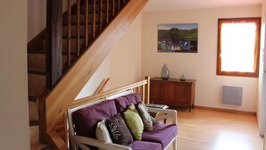 Un salon avec un escalier en bois, un canapé avec des coussins, un tableau accroché au mur et une fenêtre avec des stores.