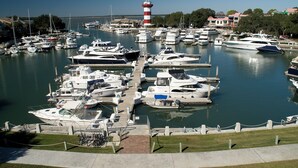 Un port de plaisance où sont amarrés de nombreux bateaux, un phare en arrière-plan et un ciel dégagé.
