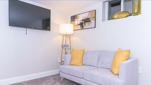 Een woonkamer met een grijze bank, gele kussens, een flatscreen televisie, een vloerlamp en een schilderij aan de muur.