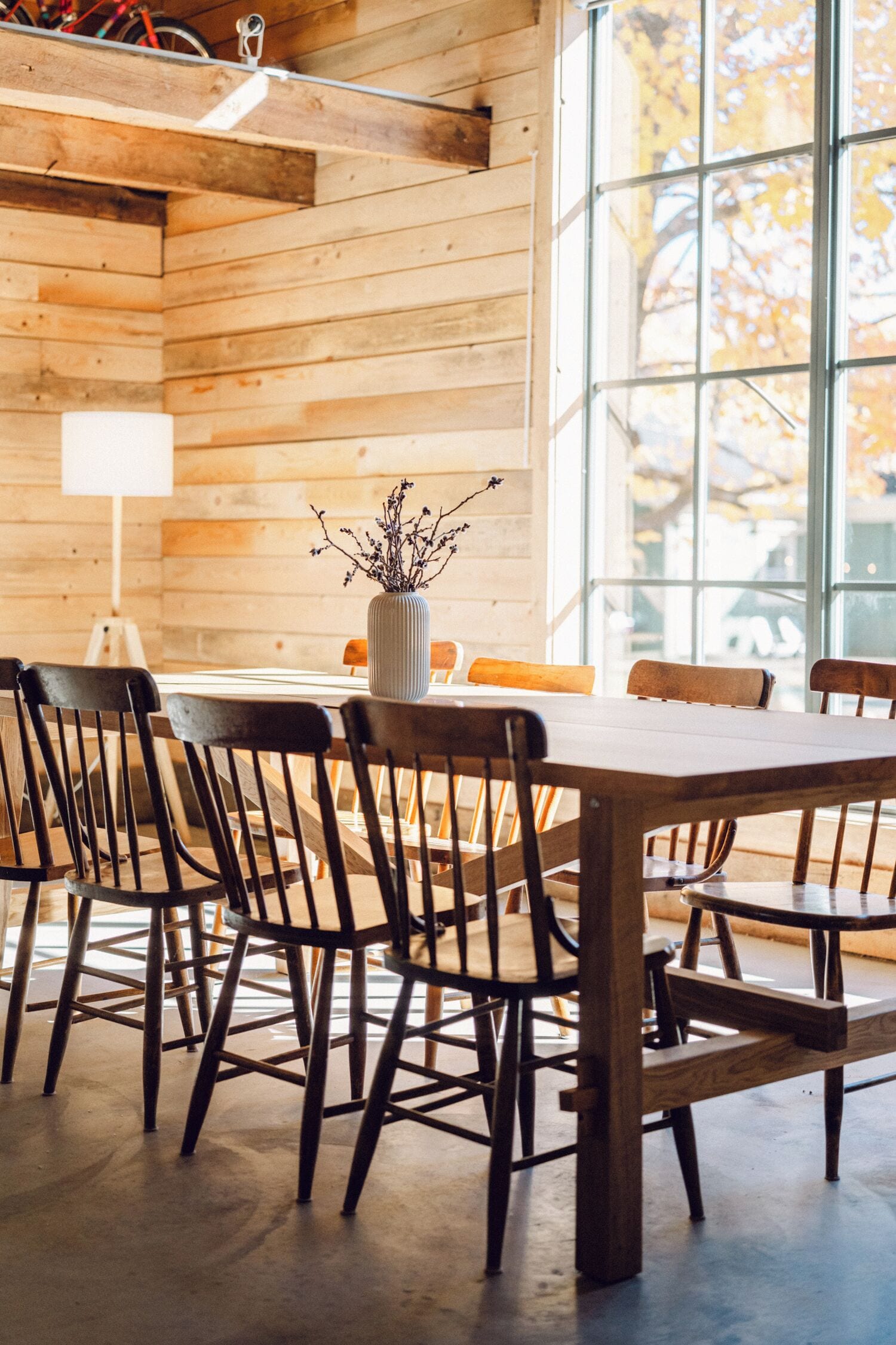 A wooden interior with a dining table and chairs, a vase with branches, and large windows.