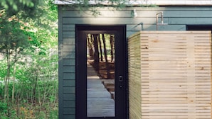 Una terraza de madera que conduce a una cabaña pequeña con una puerta oscura, rodeada de árboles.