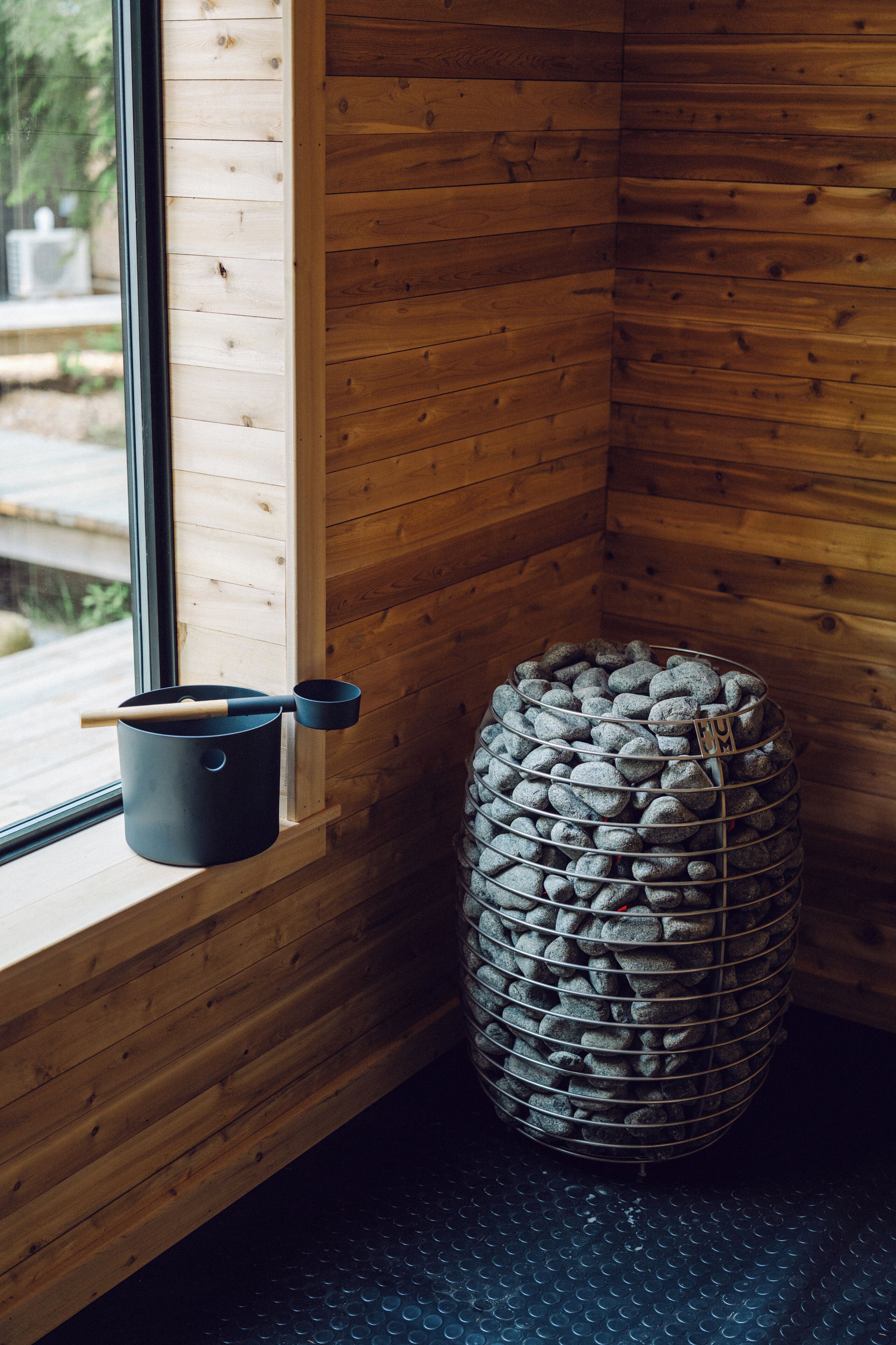 A corner of a room with wooden walls, a window, a black pot with a wooden handle, and a wire basket filled with rocks.