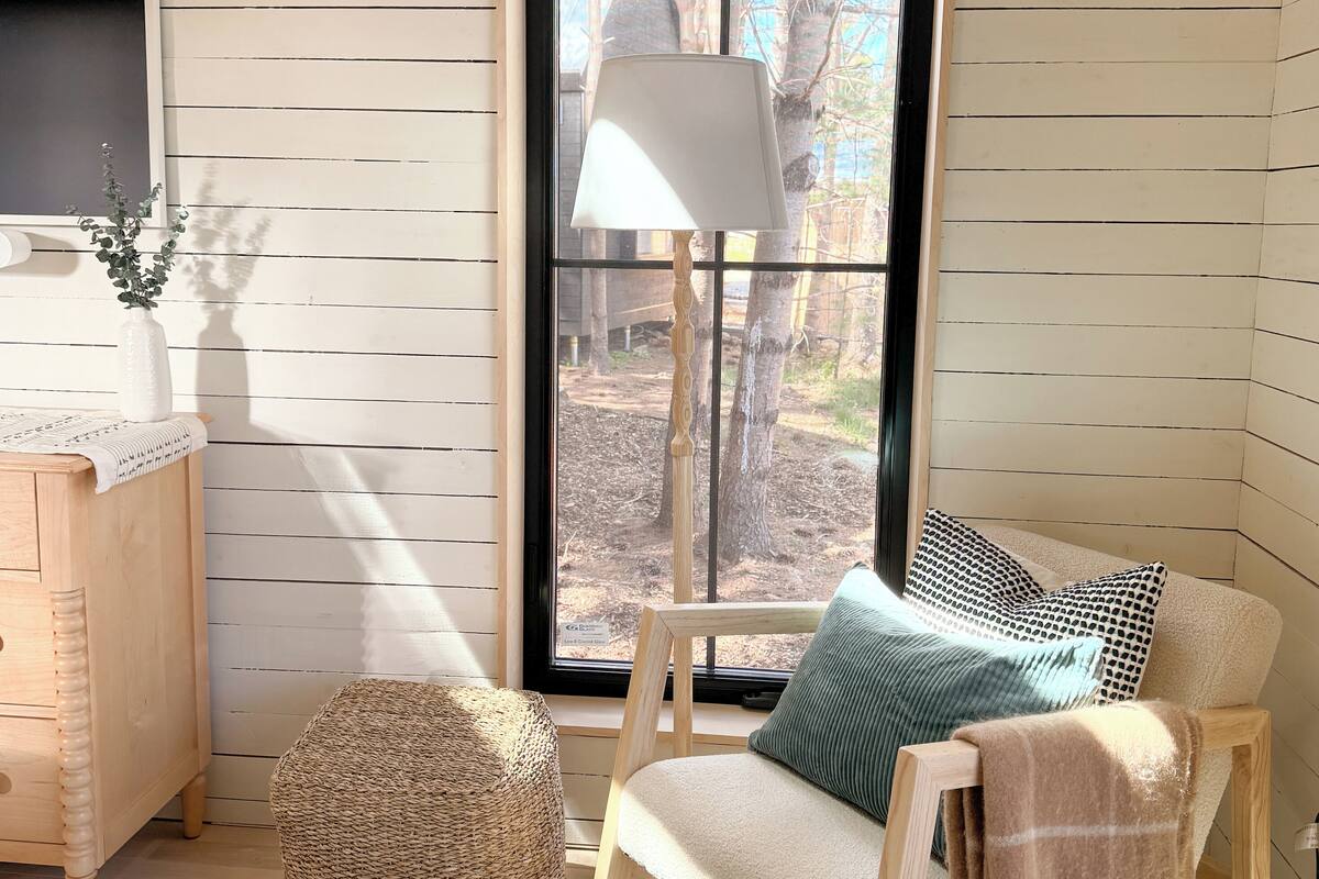A cozy room with a wooden floor, a woven pouf, a wooden chair with a blanket, a lamp, and a view of trees through a window.