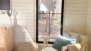 Una habitación acogedora con suelo de madera, un pouf tejido, una silla de madera con una manta, una lámpara y vistas a árboles a través de una ventana.