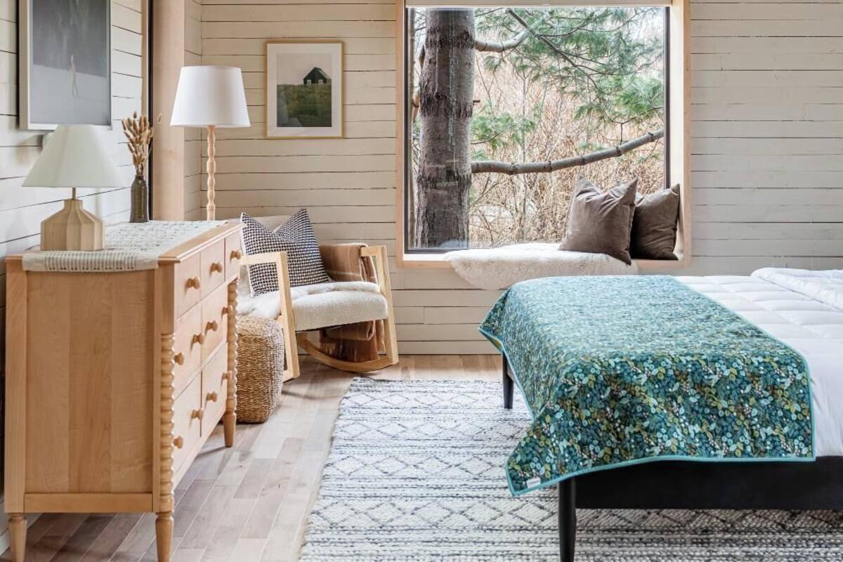 A cozy bedroom with a sloped ceiling, wooden walls, a large window with a view of trees, a bed with a floral bedspread, a bedside table with a lamp, and a dresser with drawers.