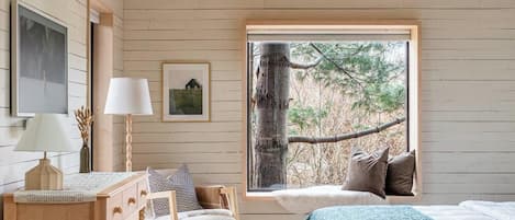 A cozy bedroom with a sloped ceiling, wooden walls, a large window with a view of trees, a bed with a floral bedspread, a bedside table with a lamp, and a dresser with drawers.