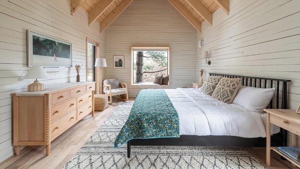 Un dormitorio con techo de madera, una cama con ropa de cama blanca, una mesita de noche, un aparador, una silla, una ventana con vistas a árboles y una alfombra estampada.