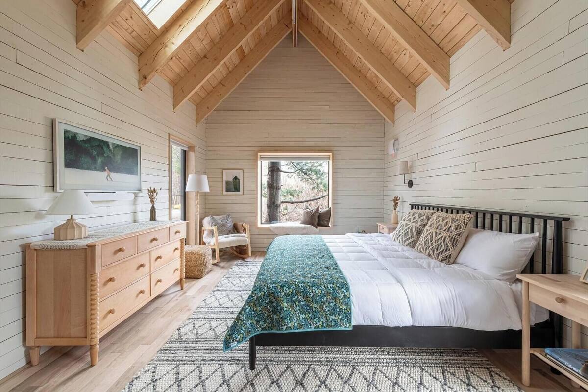 A bedroom with a wooden ceiling, a bed with white bedding, a nightstand, a dresser, a chair, a window with a view of trees, and a patterned rug.