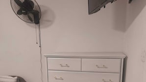 A white dresser with multiple drawers, a black fan on the wall, and a television above it.