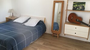 Ein Schlafzimmer mit einem Bett, einem Nachttisch mit Lampe, einer Kommode mit einer Gitarre und einem auf einem Podest stehenden Spiegel.