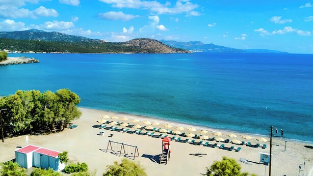 A beach with rows of sunbeds and umbrellas, a lifeguard tower, and a clear view of the sea and mountains in the background.