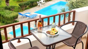 A balcony with a glass table, two chairs, a bottle of wine, glasses, a plate with food, and a view of a swimming pool and garden.