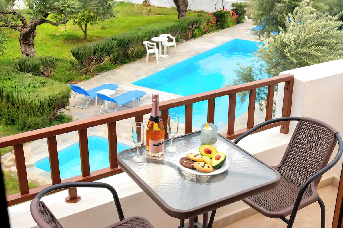 A balcony with a glass table, two chairs, a bottle of wine, glasses, a plate with food, and a view of a swimming pool and garden.
