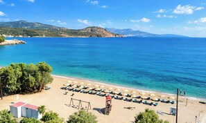 A beach with clear blue water, a row of sun loungers, and a lifeguard tower. - Plaisir Beachside Apts (Agios Nikolaos)