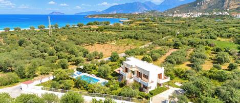 A modern villa with a pool, surrounded by lush greenery and a view of the sea and mountains.