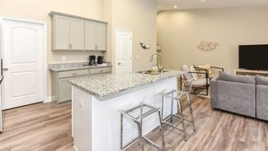 Una cocina moderna con encimeras de granito, una isla central con taburetes de bar y un comedor con un sofá gris y un televisor de pantalla plana.