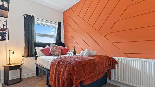 Une chambre avec un lit, une table de chevet, une fenêtre et un mur d’accent de couleur orange.