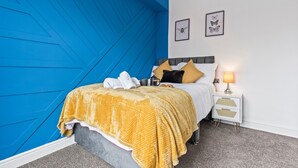 Une chambre avec un mur d’accent bleu, un lit recouvert d’une couverture jaune, deux oreillers, une table de chevet avec une lampe et deux tableaux encadrés accrochés au mur.