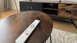 A modern living room with a round wooden coffee table, a remote control, a TV displaying the Netflix logo, a floor lamp, and a wooden cabinet with two candles. - Facing the castle & Air-conditioned ~ El Castillo ~ (Surgères)