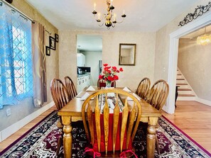 A dining room with a wooden table, six chairs, a chandelier, a vase with red flowers, and a patterned rug.