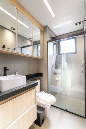 A modern bathroom with a large mirror, a white sink, a toilet, and a glass-enclosed shower area. - Residencial Pacobá (São Paulo)